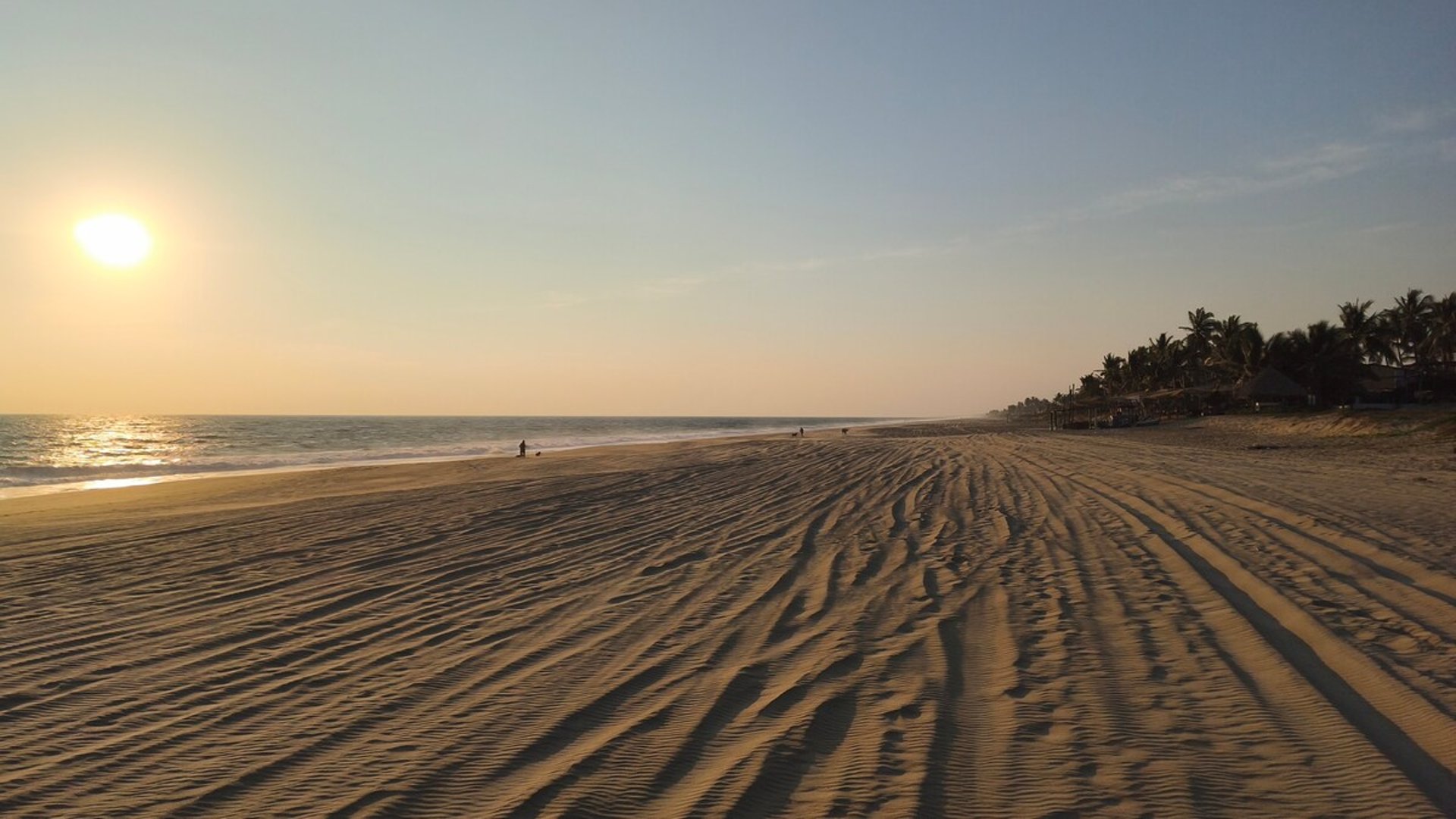 Amplia playa de arena con huellas al atardecer, mar tranquilo y palmeras al fondo bajo luz dorada