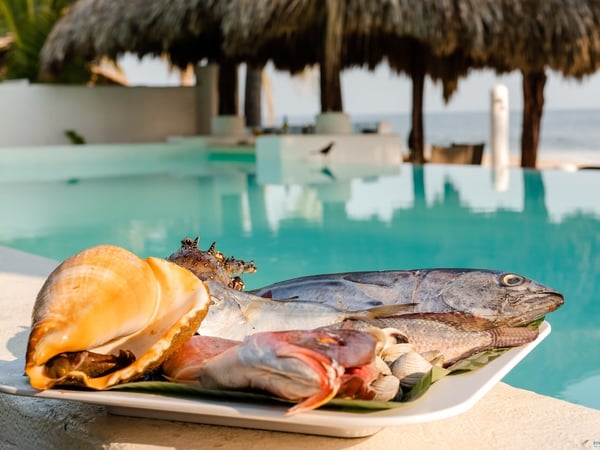 Charola de mariscos y pescado fresco en el borde de la alberca con palapa y mar al fondo