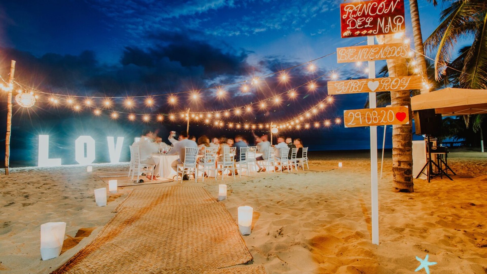 Boda en la playa al anochecer con luces festivas, letras LOVE iluminadas y mesas decoradas junto al mar