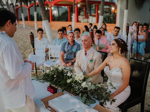 Ceremonia de boda en playa bajo palapa naranja con antorchas tiki y sillas blancas frente al mar