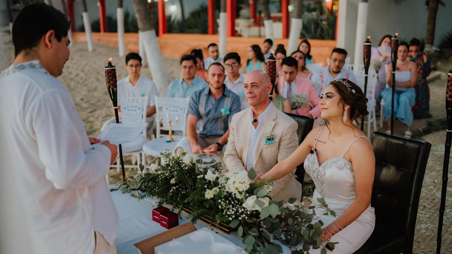 Ceremonia de boda en playa bajo palapa naranja con antorchas tiki y sillas blancas frente al mar