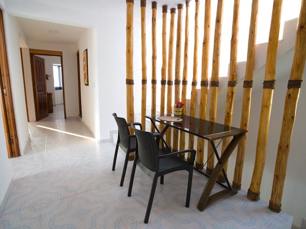 Área de comedor interior con mesa de vidrio y sillas negras, delimitada por una decorativa celosía de troncos de bambú atados, pasillo al fondo