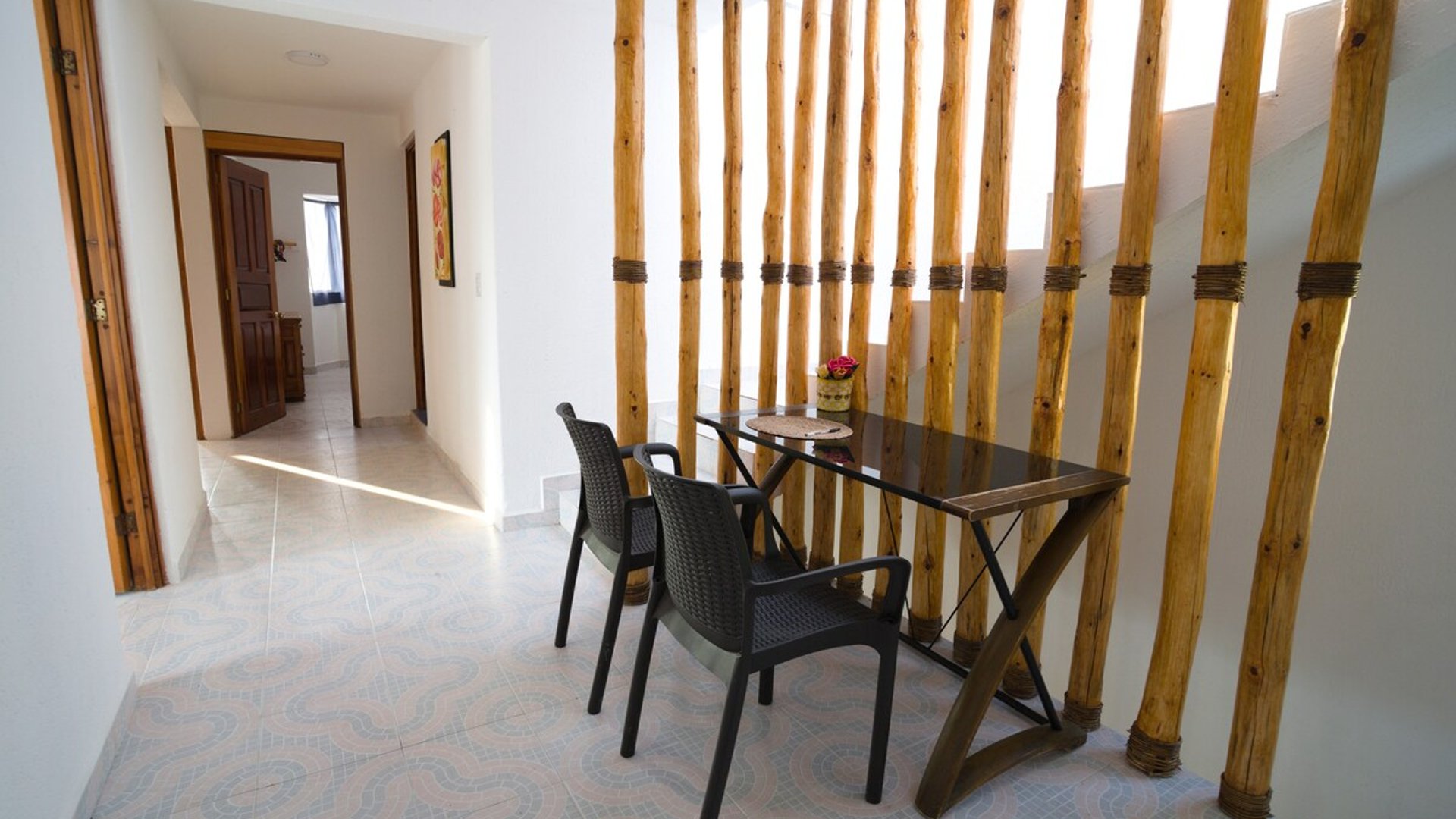 Área de comedor interior con mesa de vidrio y sillas negras, delimitada por una decorativa celosía de troncos de bambú atados, pasillo al fondo