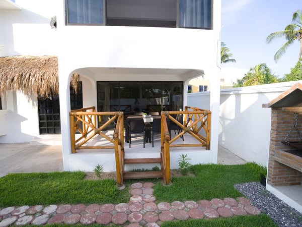 Terraza exterior con barandal de bambú, comedor y asador de ladrillo junto a jardín y palapa en villa blanca
