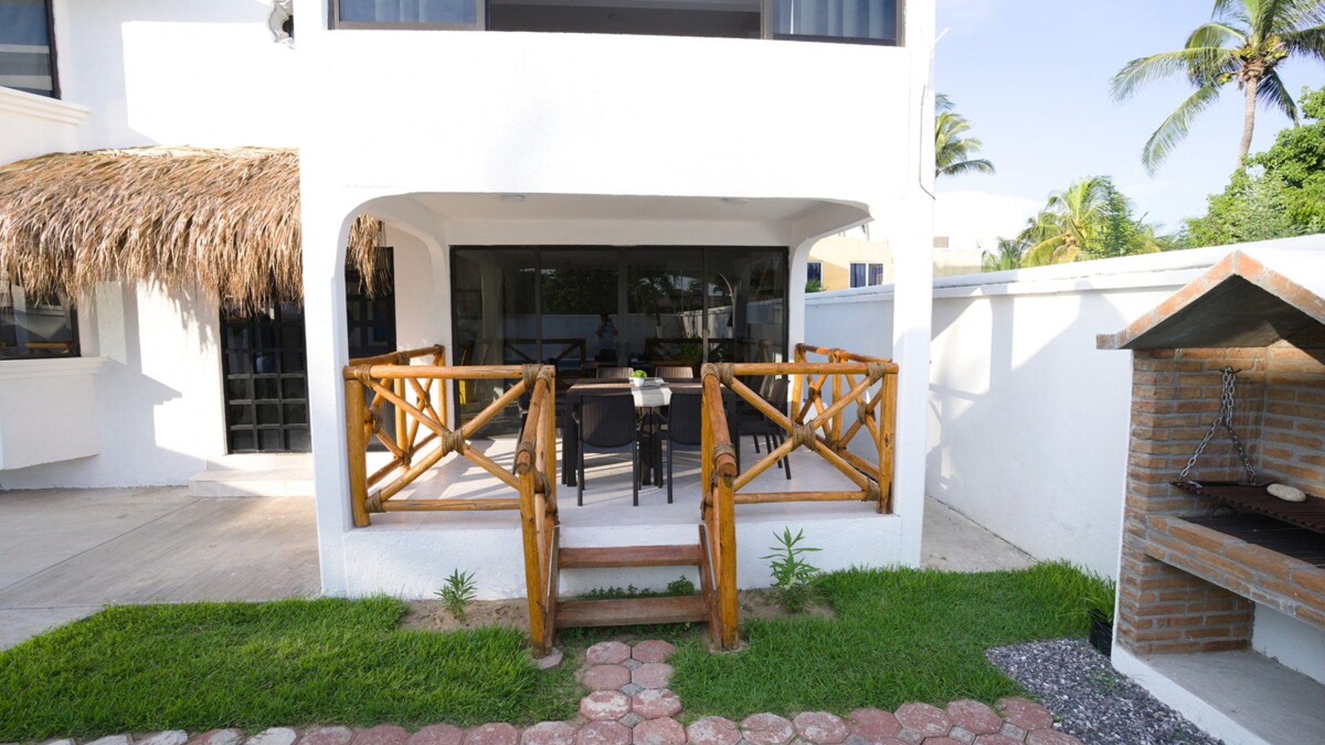 Terraza exterior con barandal de bambú, comedor y asador de ladrillo junto a jardín y palapa en villa blanca