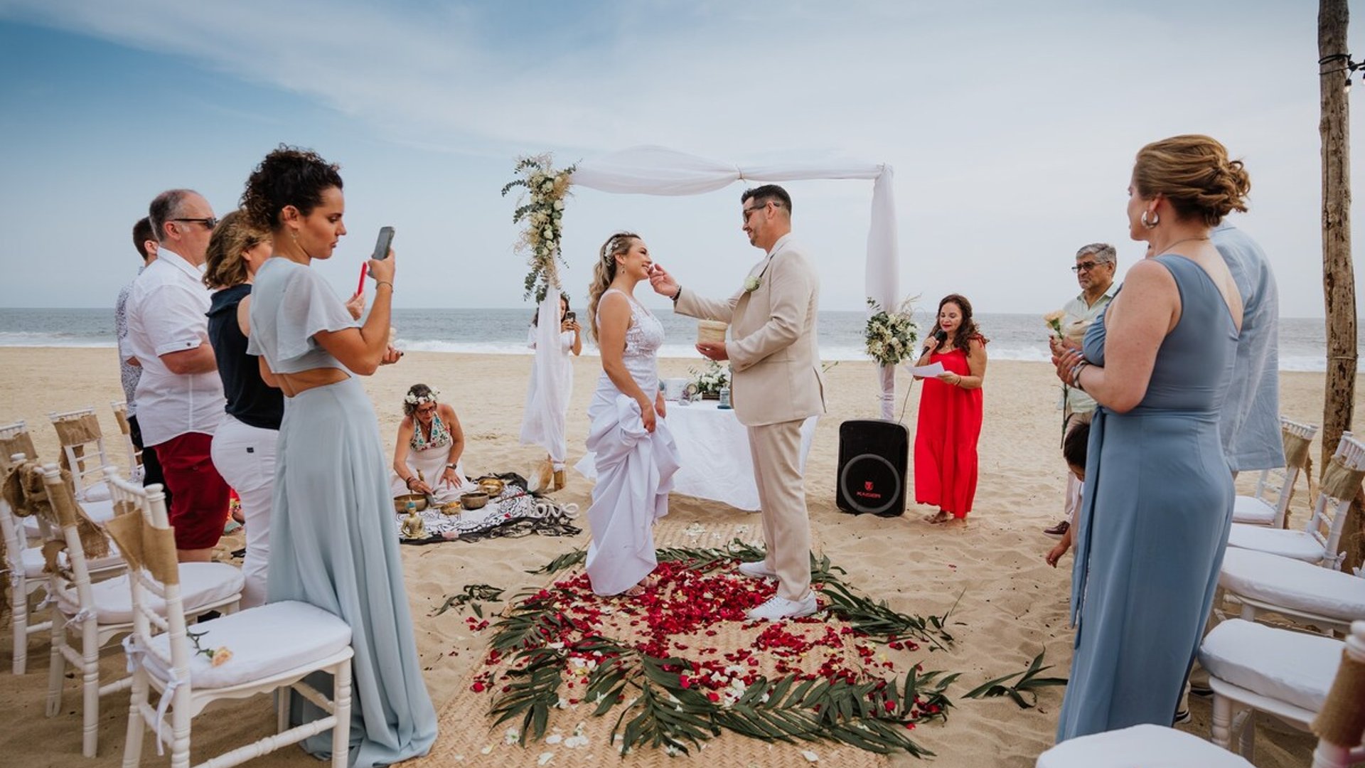 Ceremonia de boda en la playa con arco floral blanco, pétalos de rosas y mar al fondo