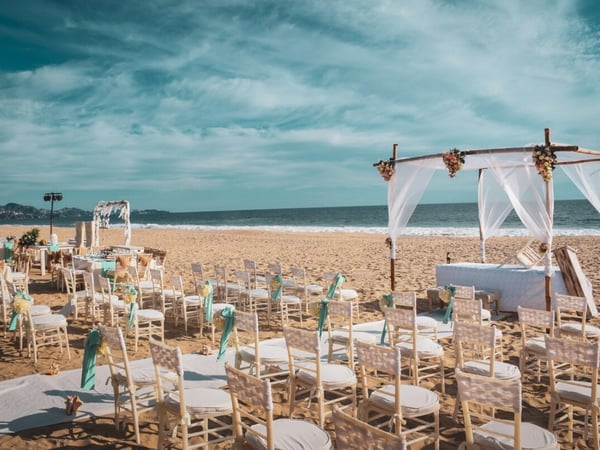Ceremonia de boda montada en la playa con altar de bambú, telas blancas y flores, sillas Tiffany con listones turquesa frente al mar