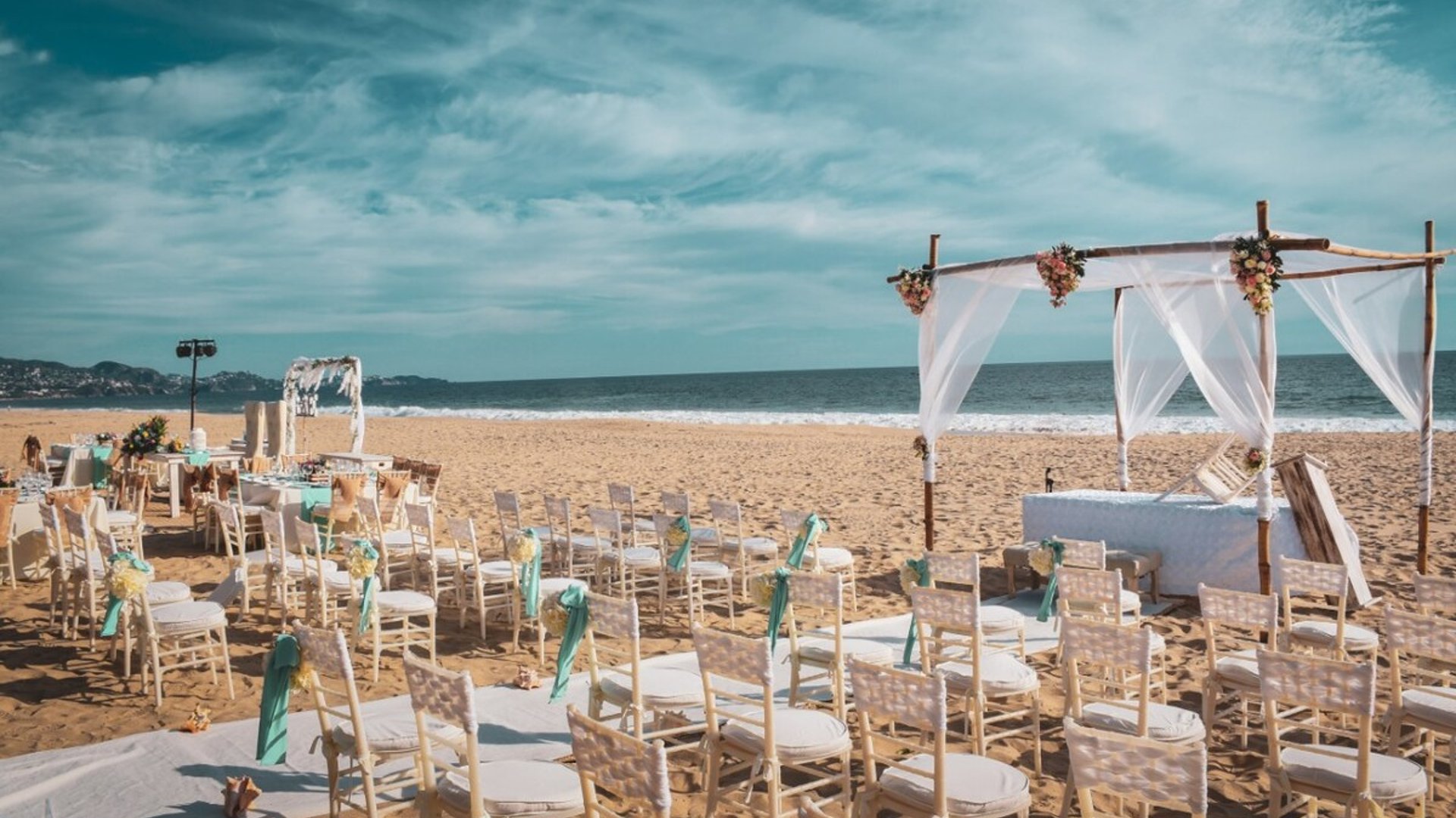 Ceremonia de boda montada en la playa con altar de bambú, telas blancas y flores, sillas Tiffany con listones turquesa frente al mar