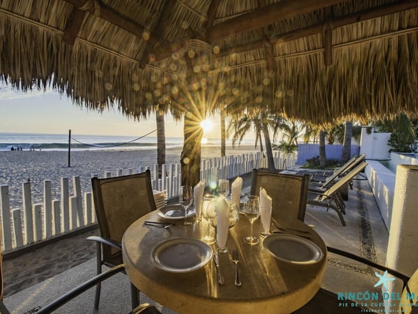 Mesa para dos bajo palapa frente al mar al atardecer, con vajilla elegante y vista directa a la playa