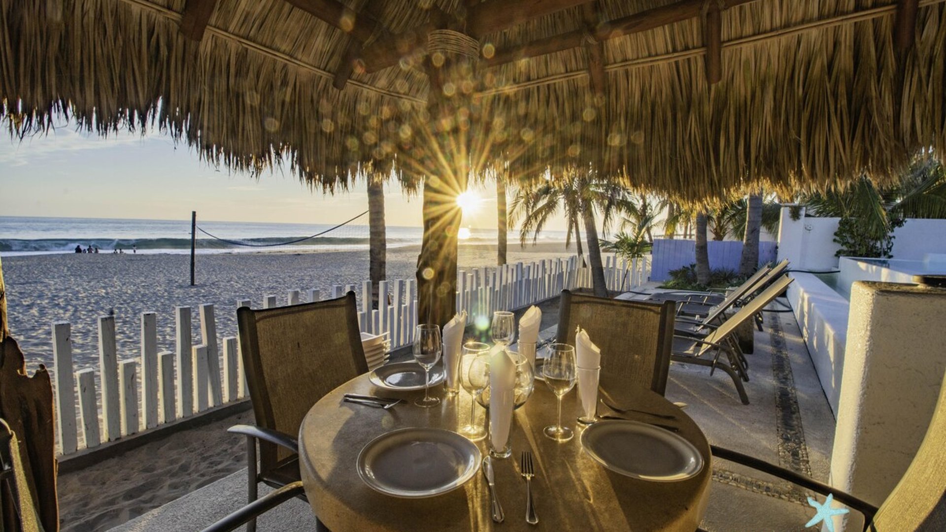 Mesa para dos bajo palapa frente al mar al atardecer, con vajilla elegante y vista directa a la playa