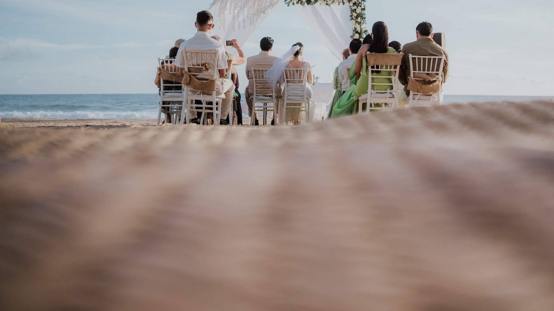 Ceremonia de boda en playa con arco floral y cortinas blancas frente al mar con invitados