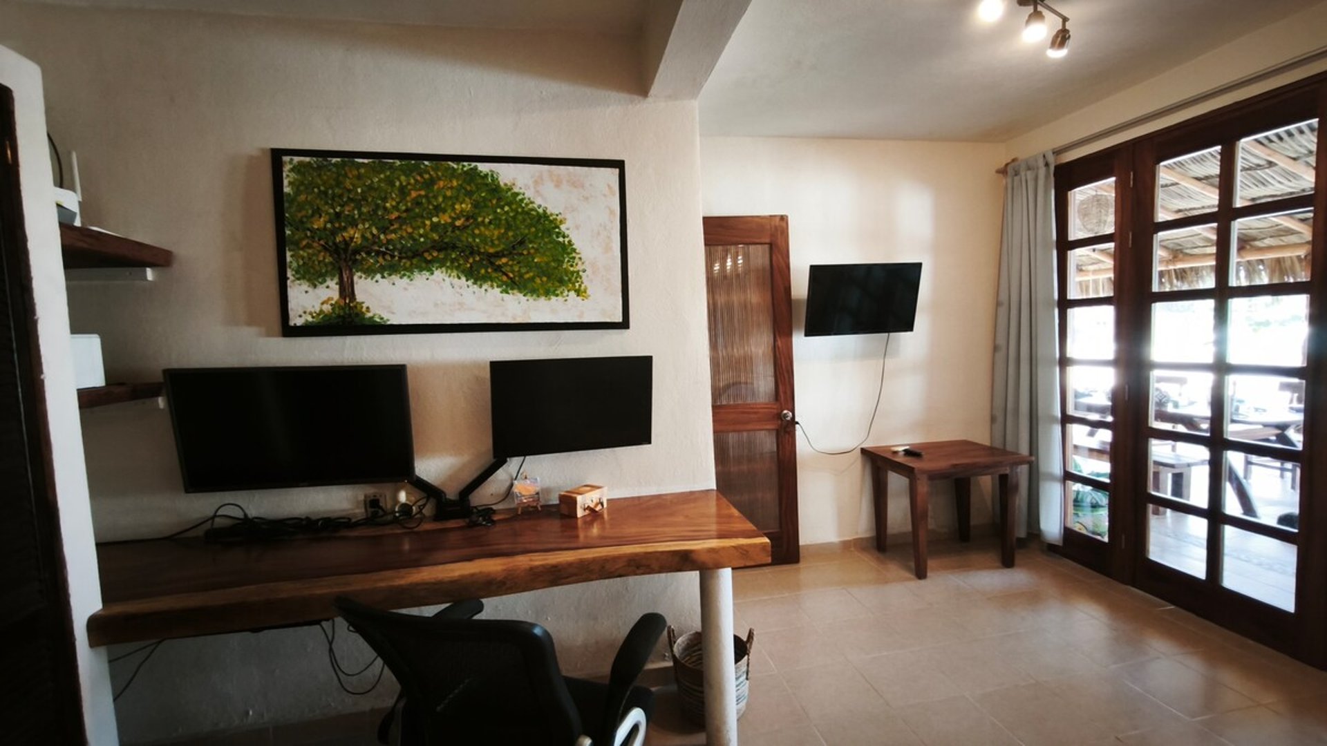 Área de trabajo con escritorio de madera maciza, dos monitores y pintura de árbol, con acceso a terraza exterior