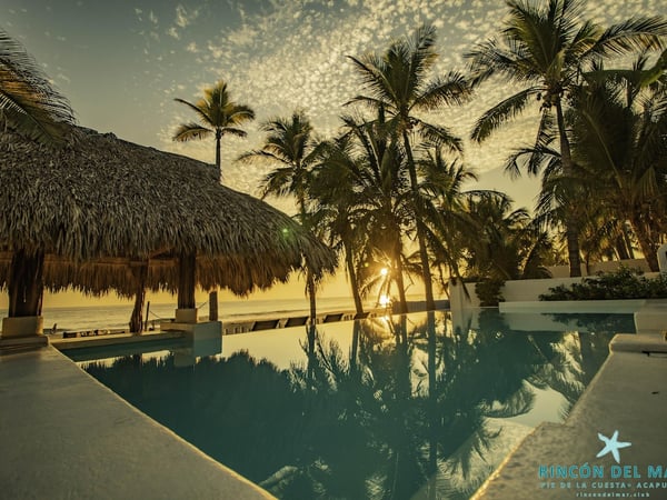 Alberca frente al mar al atardecer con palapa y palmeras reflejadas en el agua tranquila