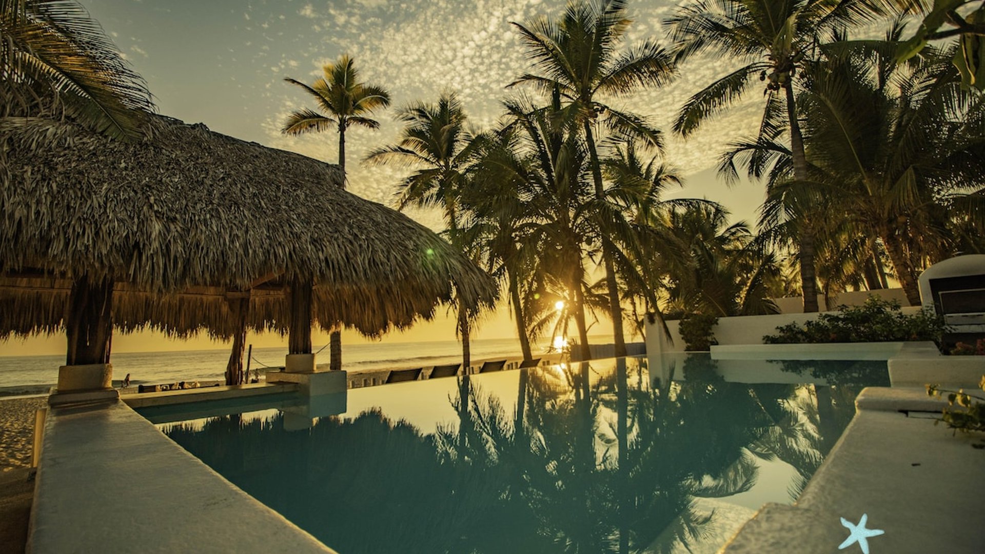 Alberca frente al mar al atardecer con palapa y palmeras reflejadas en el agua tranquila