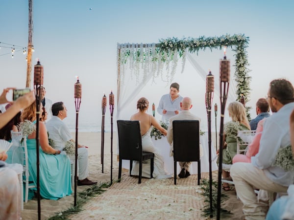 Ceremonia de boda en playa con arco floral blanco, antorchas tiki y vista al mar al atardecer