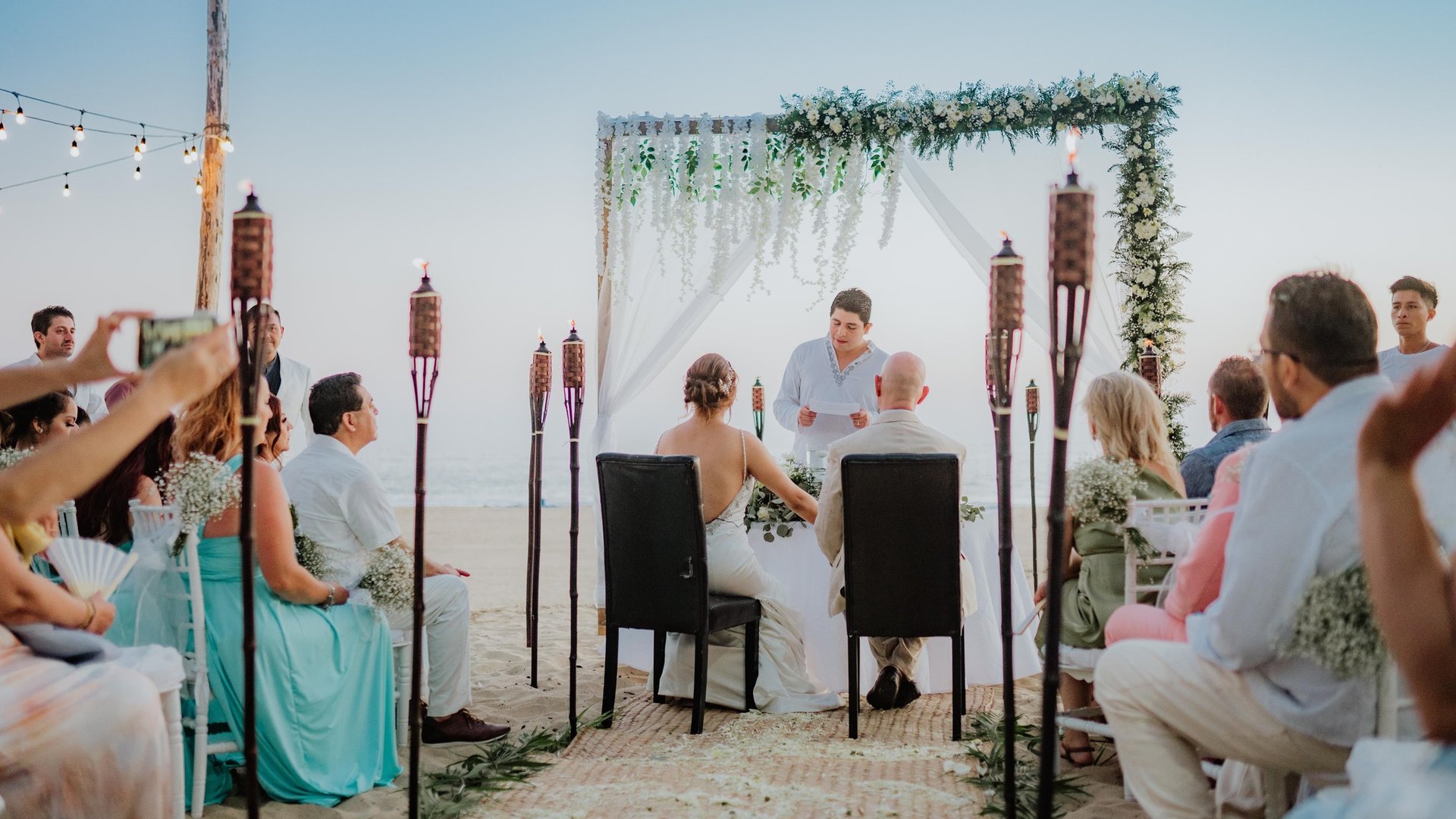Ceremonia de boda en playa con arco floral blanco, antorchas tiki y vista al mar al atardecer