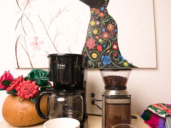 Rincón de café con cafetera, molino Cuisinart, taza y granos sobre mantel bordado colorido con cuadro decorativo al fondo
