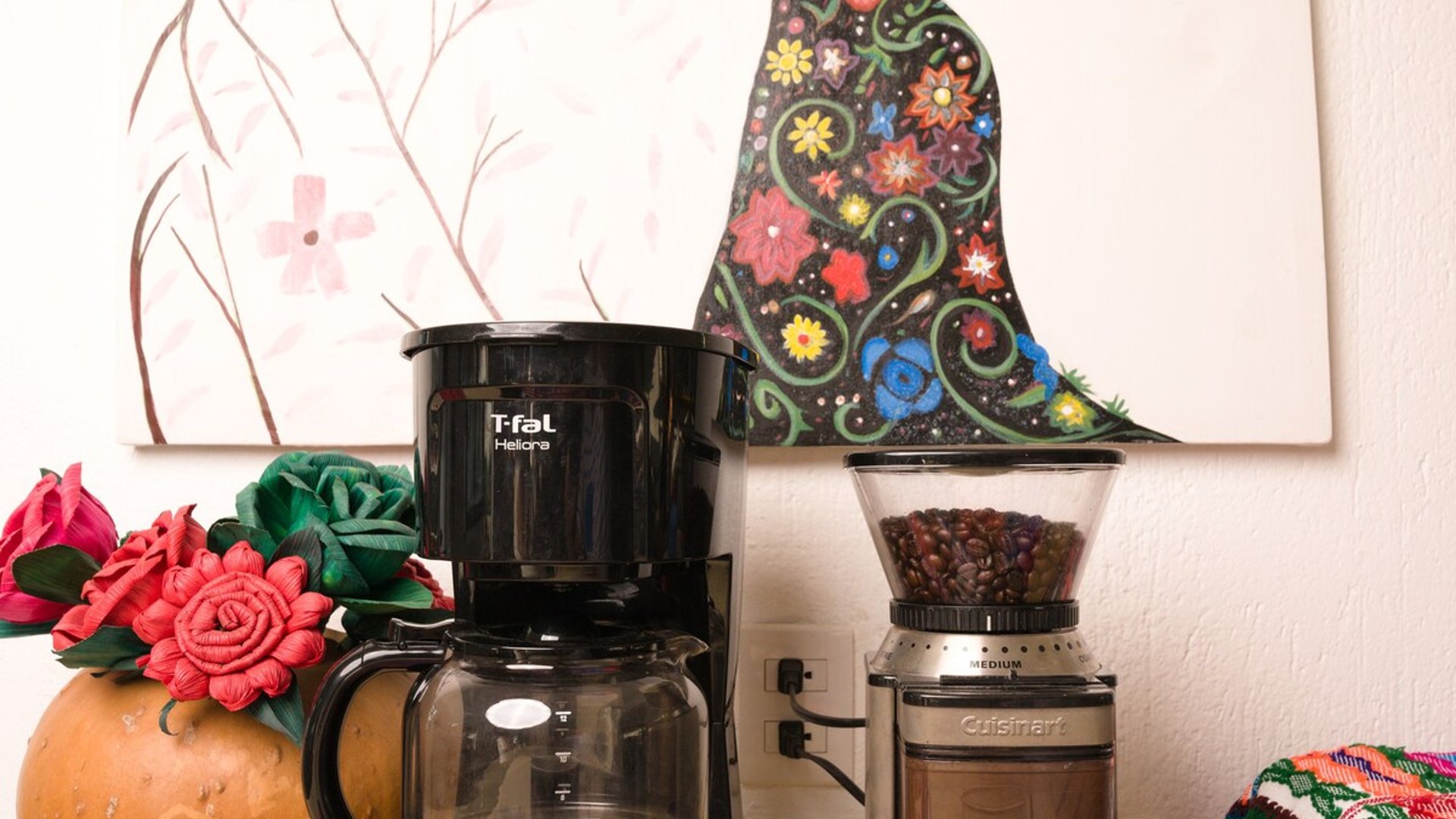 Rincón de café con cafetera, molino Cuisinart, taza y granos sobre mantel bordado colorido con cuadro decorativo al fondo