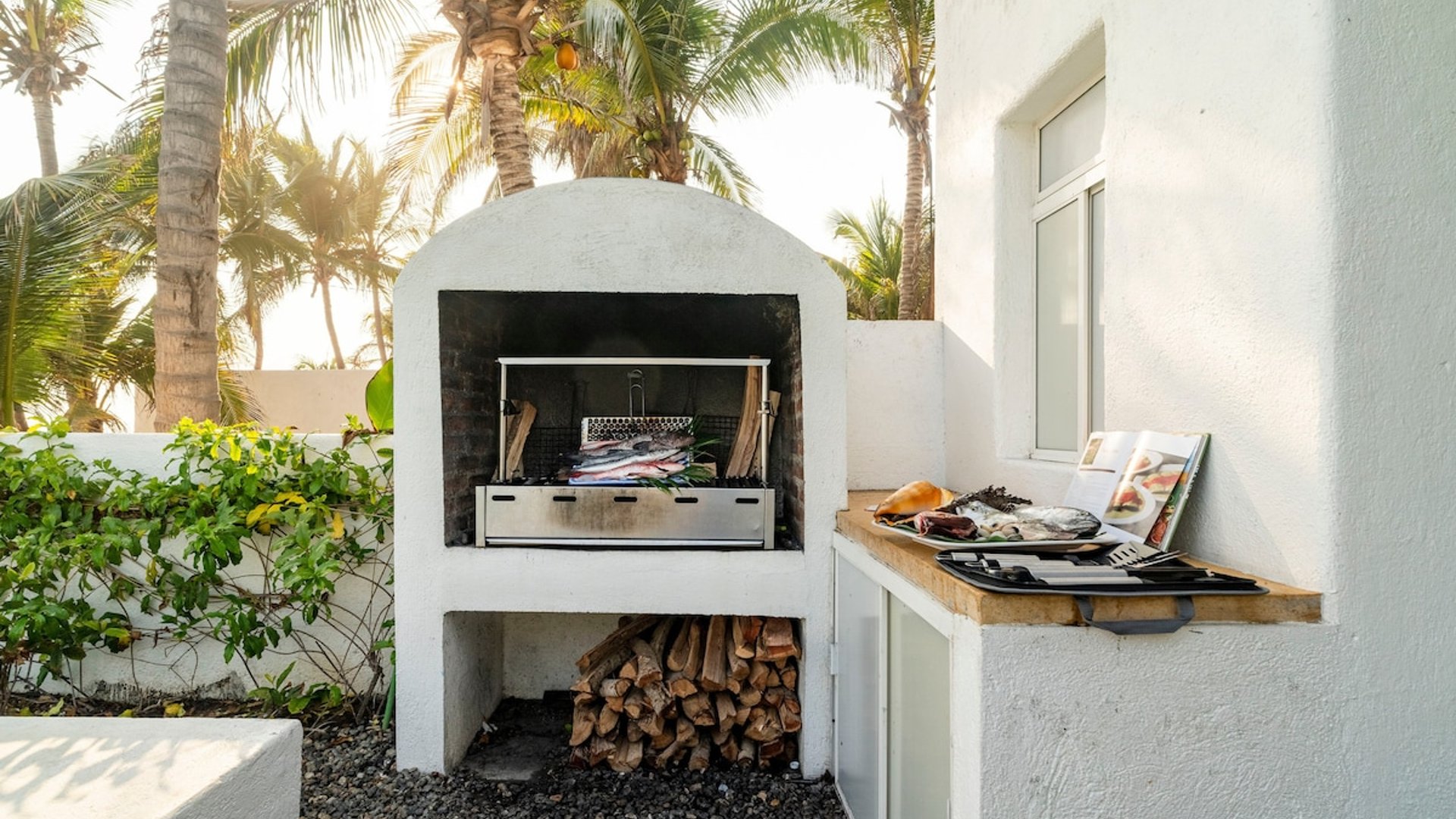 Asador de leña exterior con pescados frescos listos para cocinar, rodeado de palmeras a la luz del atardecer