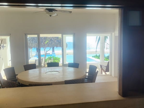 Comedor interior con mesa redonda y sillas oscuras, vistas al mar y palapa desde ventanales