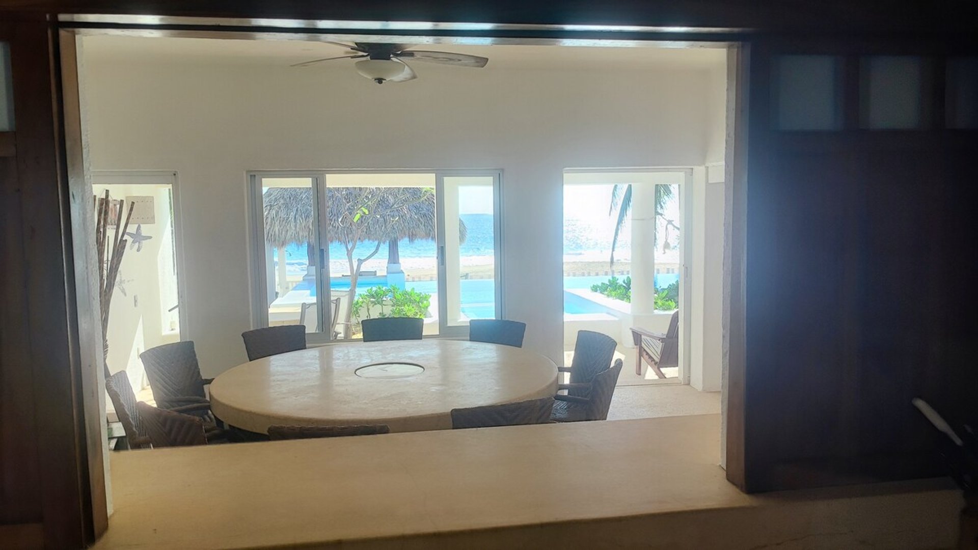 Comedor interior con mesa redonda y sillas oscuras, vistas al mar y palapa desde ventanales