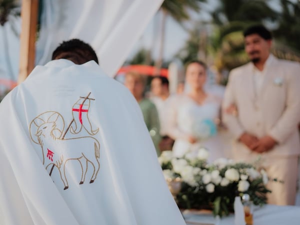 Ceremonia de boda en la playa con sacerdote, pareja de novios y arreglo floral blanco al atardecer