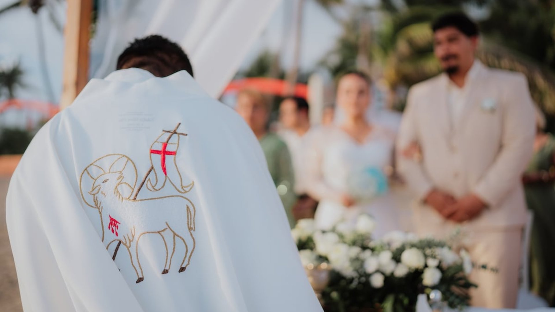 Ceremonia de boda en la playa con sacerdote, pareja de novios y arreglo floral blanco al atardecer