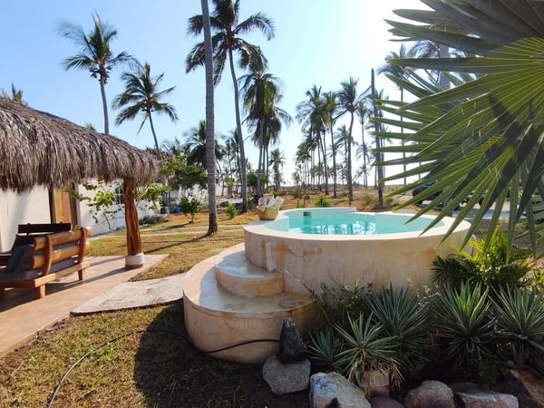 Jacuzzi redondo con escalones de piedra caliza, palapa con área lounge y palmeras al fondo bajo cielo despejado