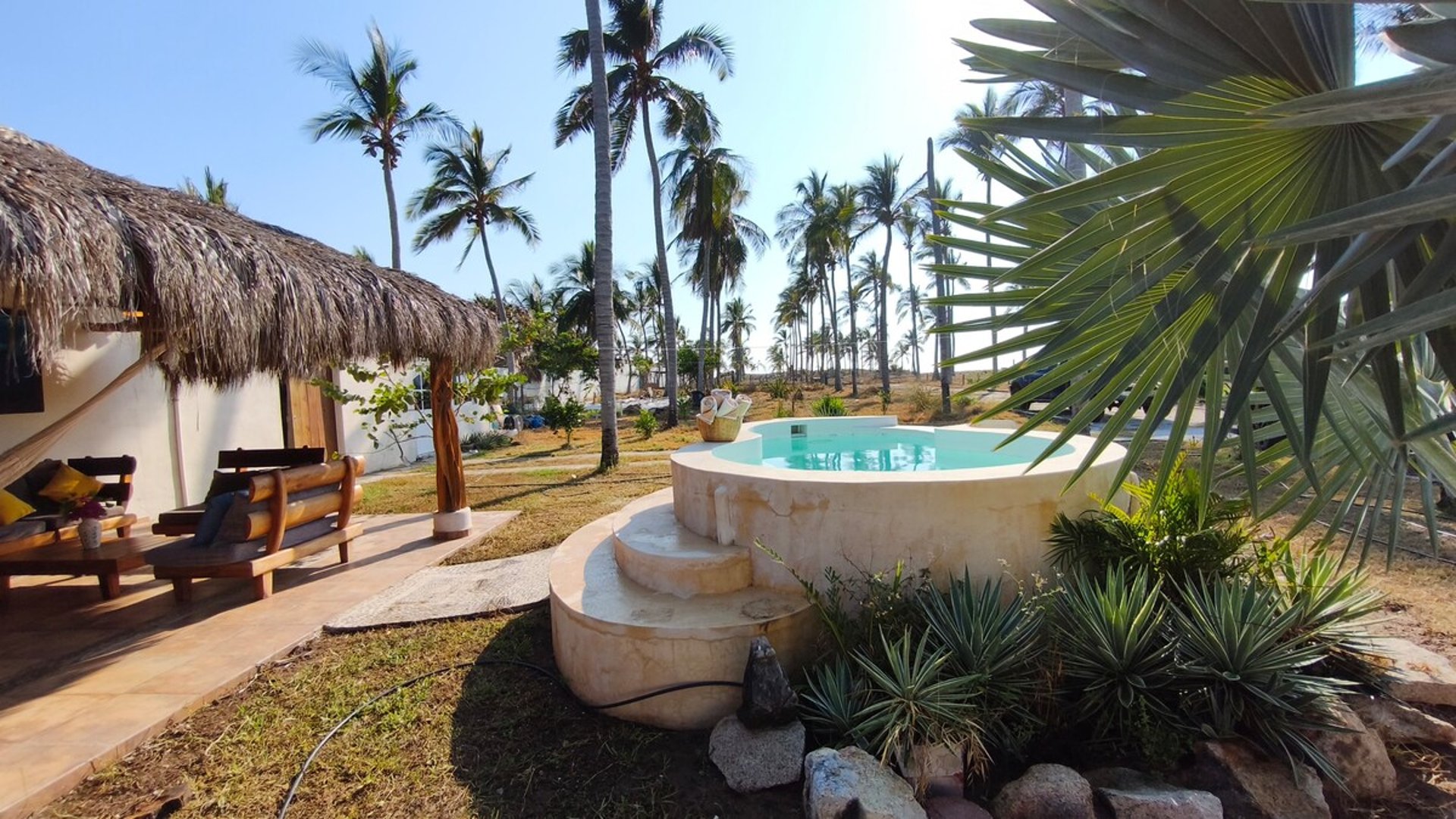 Jacuzzi redondo con escalones de piedra caliza, palapa con área lounge y palmeras al fondo bajo cielo despejado