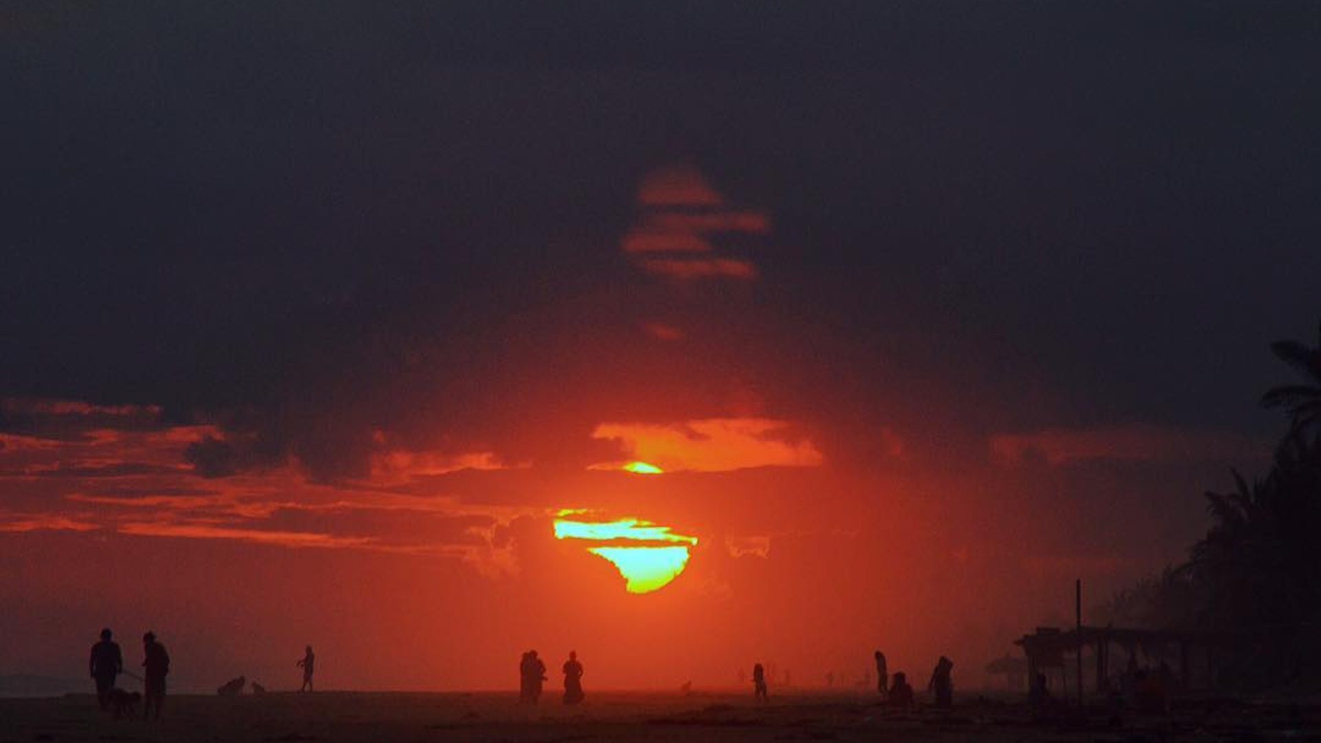 Atardecer dramático en la playa con siluetas de personas y sol entre nubes rojas