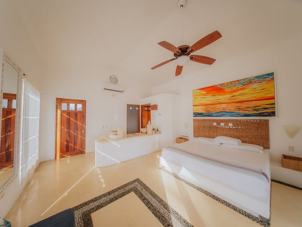 Recámara amplia con cama doble, cabecera de mimbre, cuadro de atardecer y techo alto con ventilador