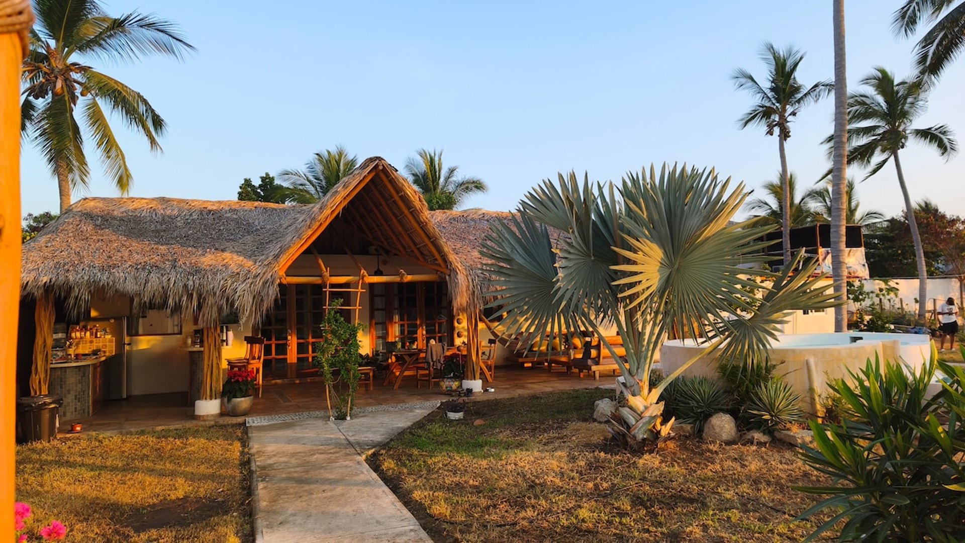 Fachada de palapa con techo de palma y estructura de bambú rodeada de palmeras y vegetación tropical al atardecer
