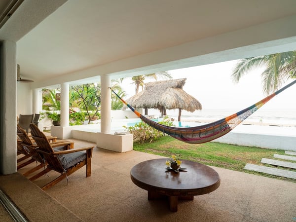 Terraza cubierta con hamaca colorida, mesa de madera y vista a palapa, alberca y mar al fondo