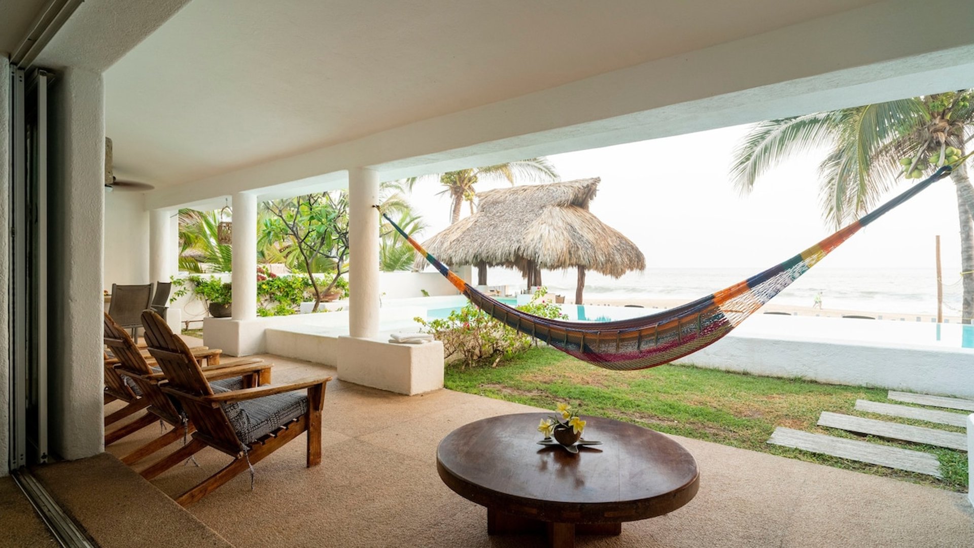 Terraza cubierta con hamaca colorida, mesa de madera y vista a palapa, alberca y mar al fondo