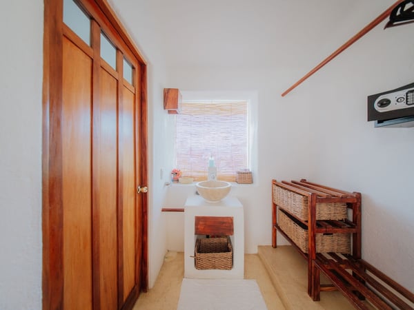 Área de lavabo tipo vessel sobre base blanca con mueble de ratán, puerta de madera y caja fuerte en recámara