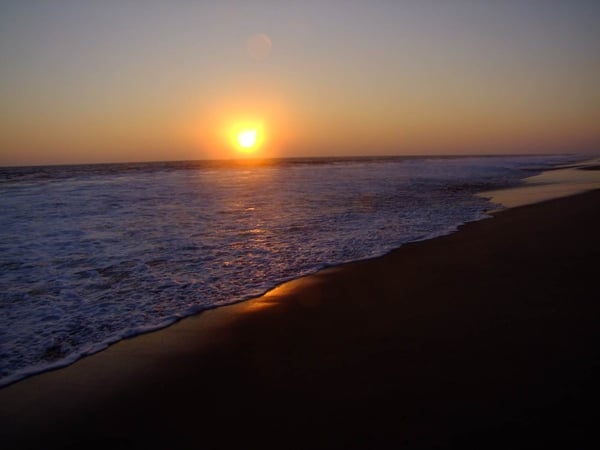 Atardecer sobre el Pacífico con sol en el horizonte, olas rompiendo en la orilla de arena oscura