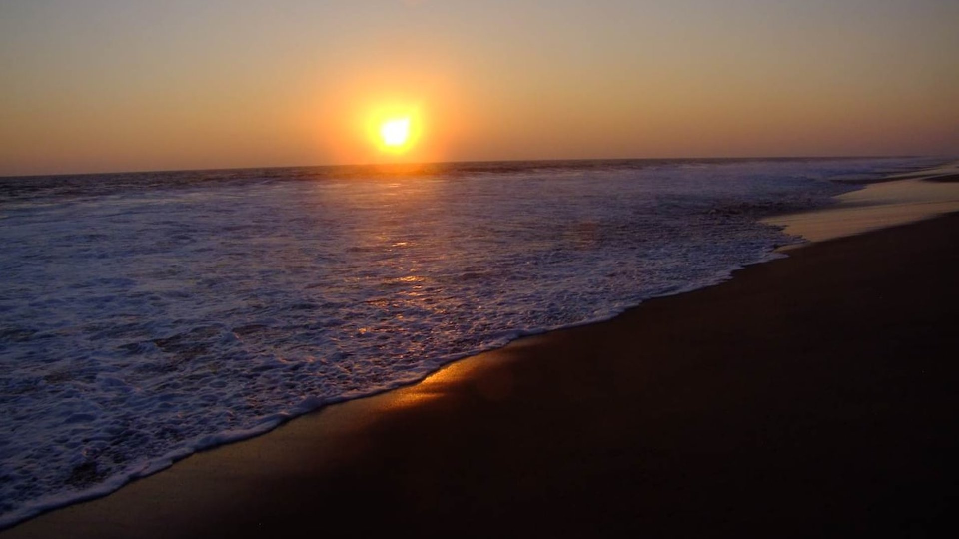 Atardecer sobre el Pacífico con sol en el horizonte, olas rompiendo en la orilla de arena oscura