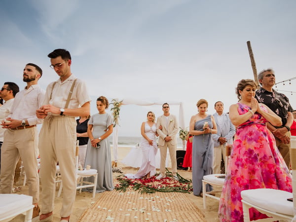 Ceremonia de boda en playa con invitados de pie frente a arco blanco y vista al mar
