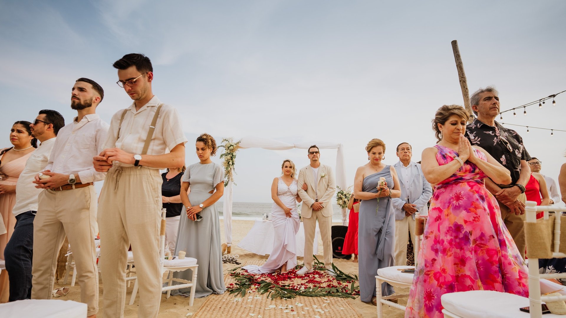 Ceremonia de boda en playa con invitados de pie frente a arco blanco y vista al mar