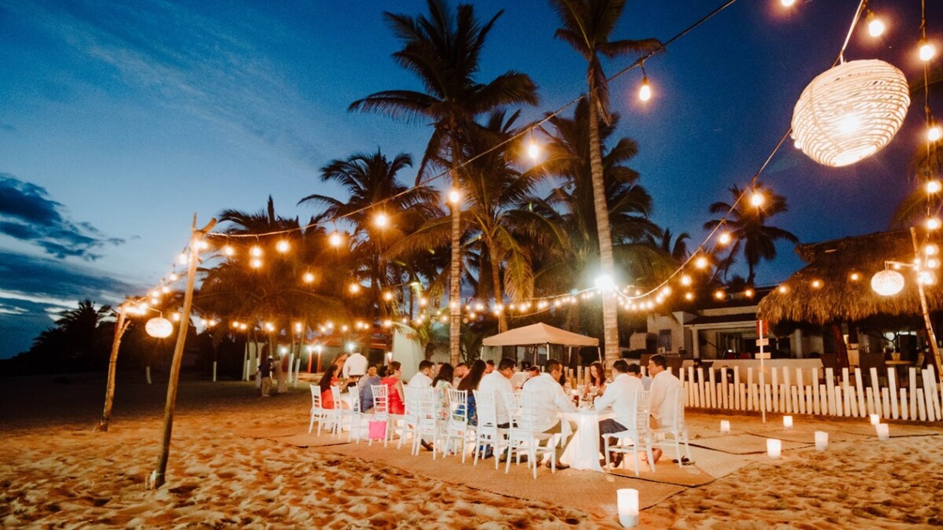 Cena de grupo en la playa al anochecer con guirnaldas de luces, palmeras y velas en la arena