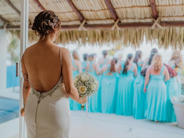 Novia con ramo de rosas blancas bajo palapa frente a damas de honor en vestidos turquesa