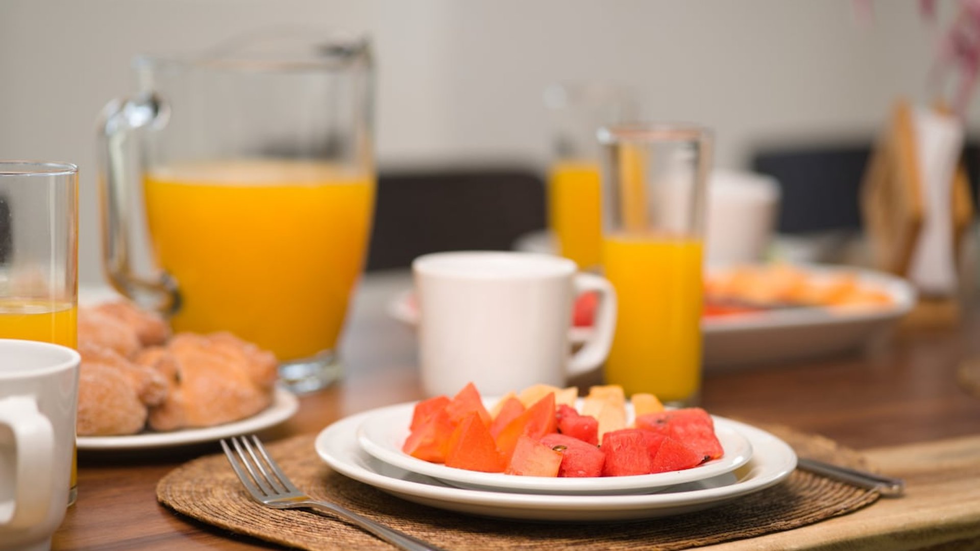 Mesa de desayuno con plato de frutas tropicales, jugo de naranja y pan dulce sobre mesa de madera