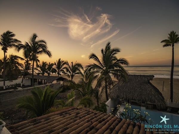 Vista aérea al atardecer de la propiedad frente al mar con palapa, alberca y palmeras en silueta