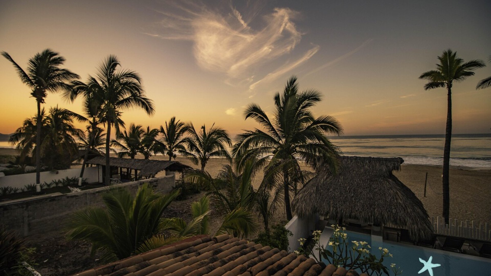 Vista aérea al atardecer de la propiedad frente al mar con palapa, alberca y palmeras en silueta