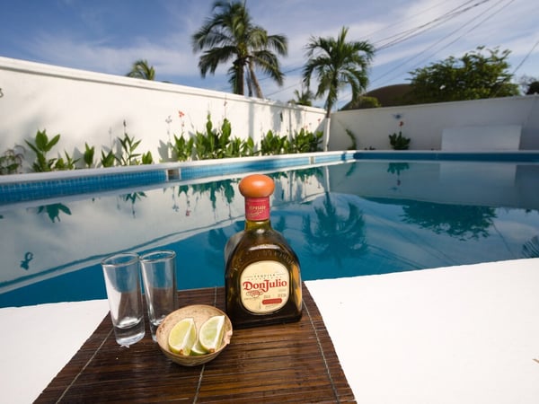 Botella de tequila con limones y copas en el borde de alberca privada rodeada de palmeras y jardín