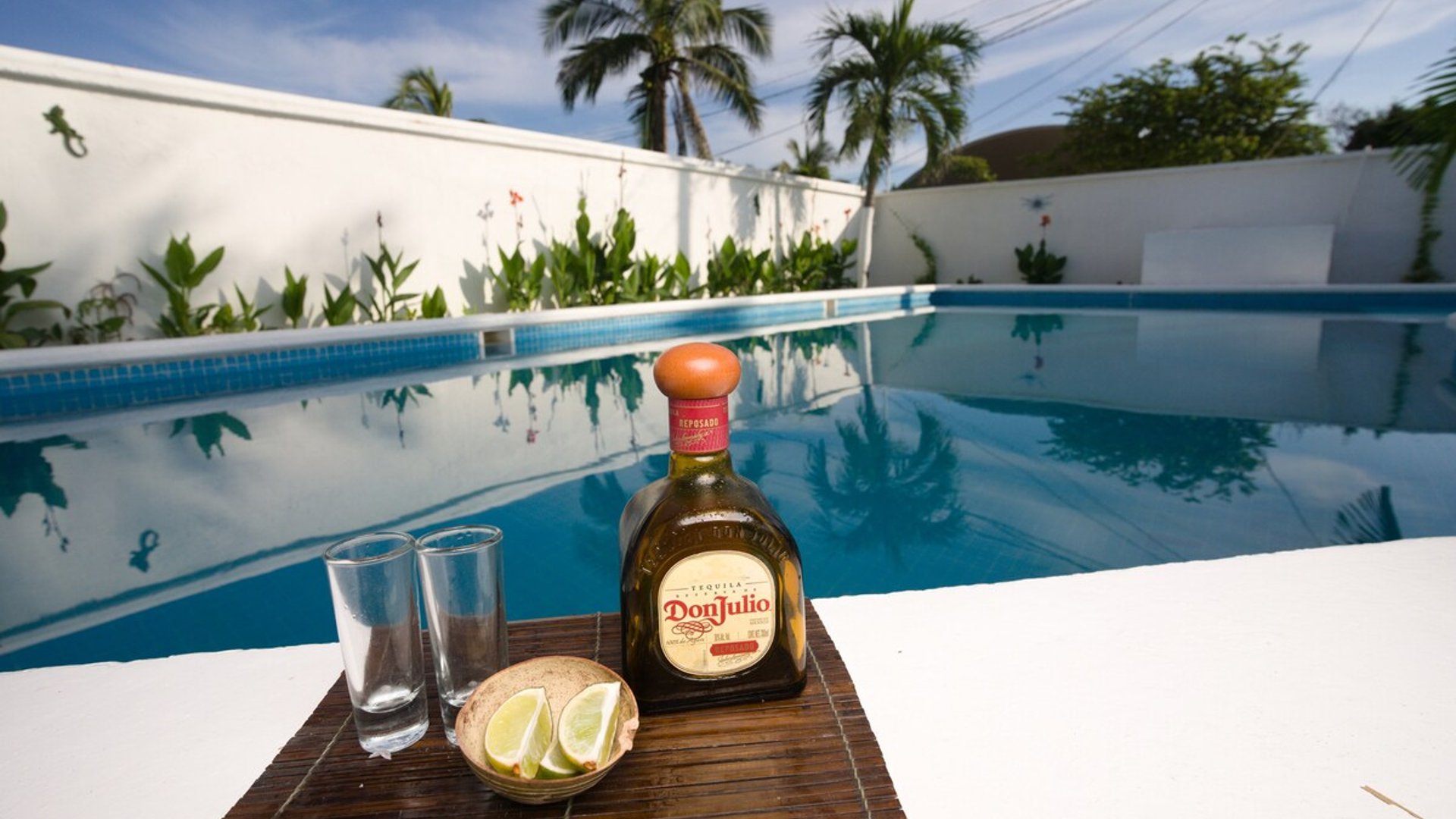 Botella de tequila con limones y copas en el borde de alberca privada rodeada de palmeras y jardín