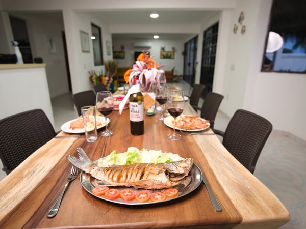 Mesa de madera larga servida con pescado a la parrilla, camarones, vino tinto y arreglo floral en comedor interior iluminado