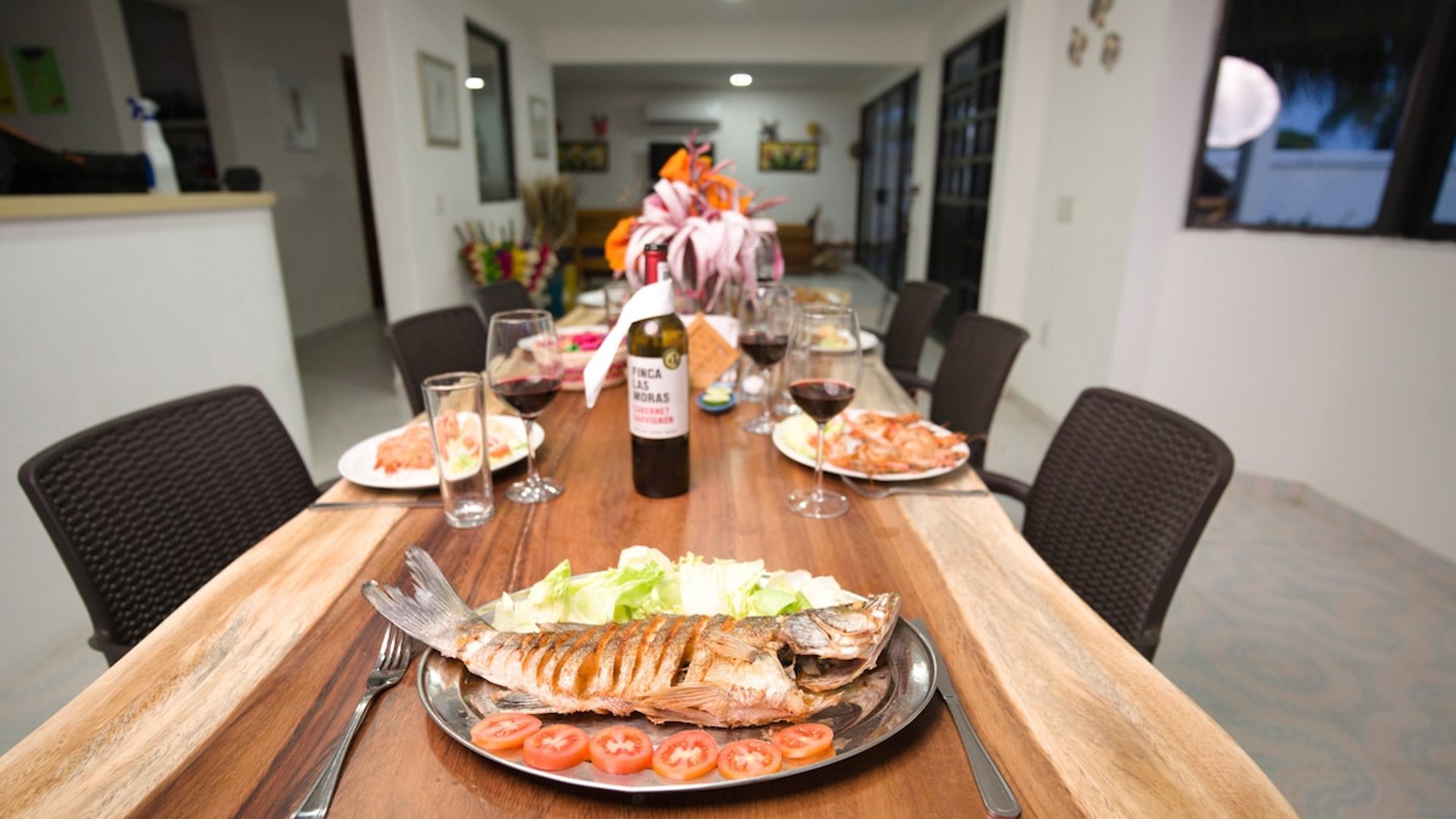 Mesa de madera larga servida con pescado a la parrilla, camarones, vino tinto y arreglo floral en comedor interior iluminado