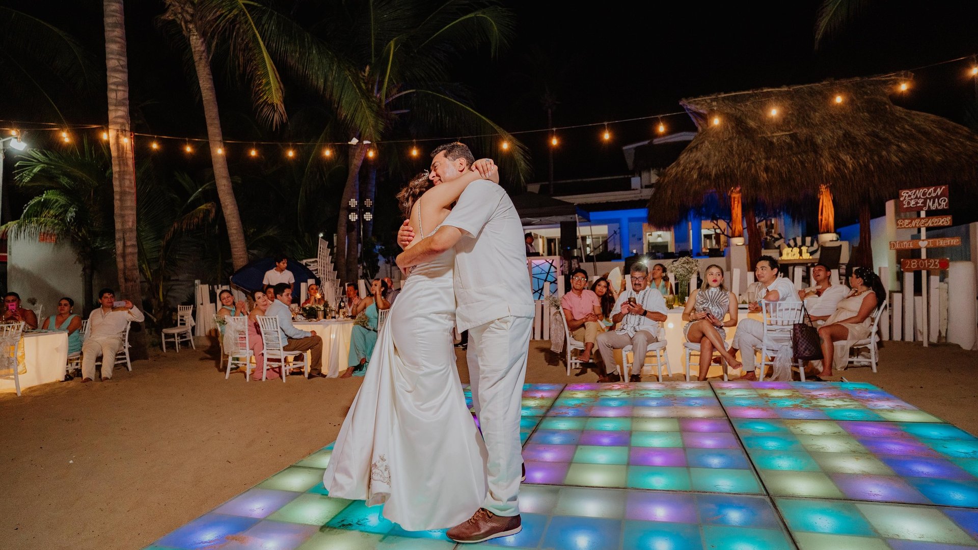 Primer baile nocturno en pista iluminada con luces de colores bajo palmeras y guirnaldas de focos