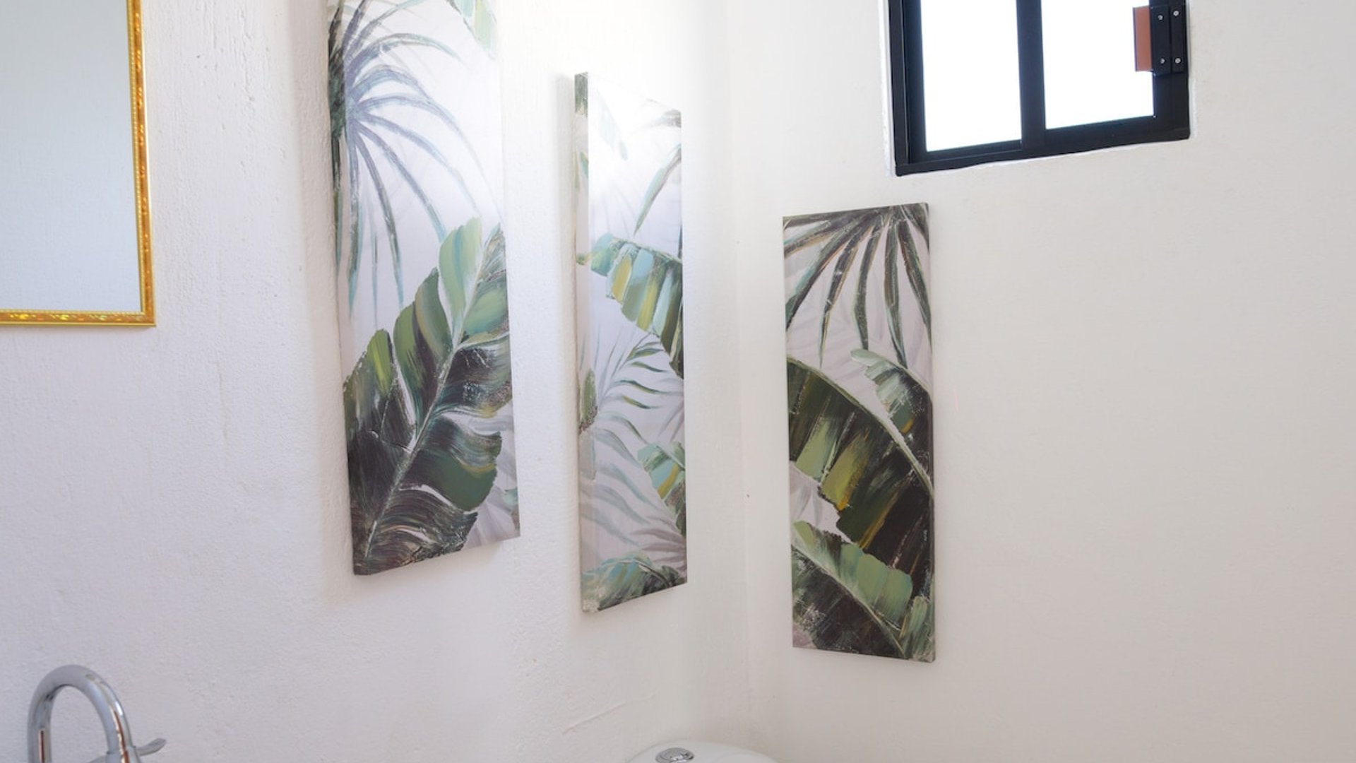 Baño blanco con lavabo, inodoro y tres cuadros tropicales de hojas verdes sobre la pared